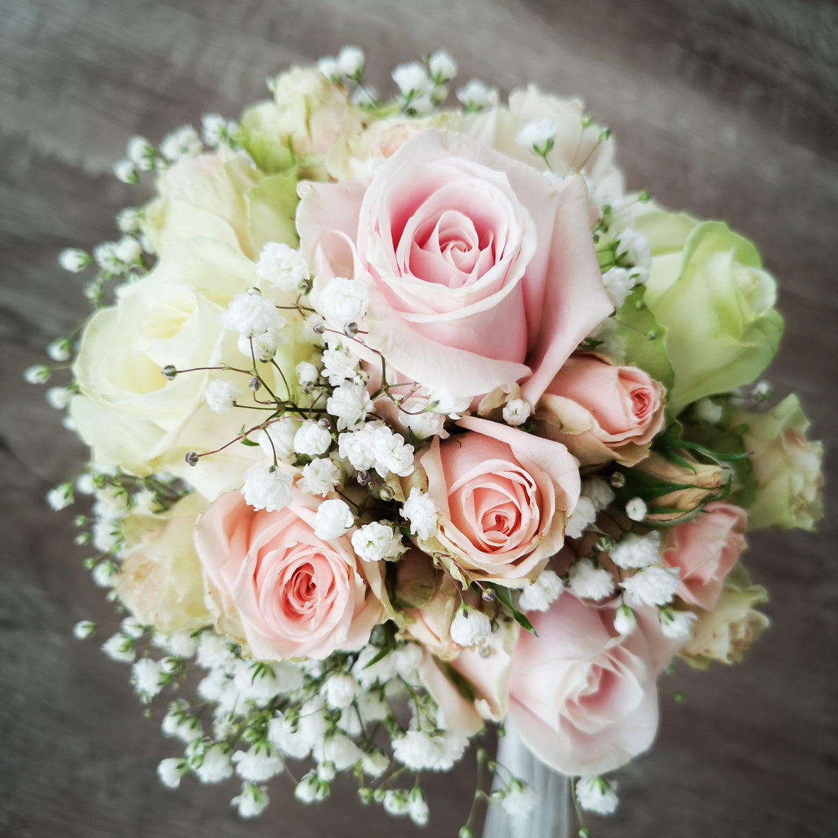 A floral bouquet featuring light pink and cream roses with white baby's breath, arranged on a rustic dark wood surface.