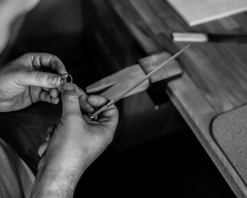 Black and white close-up of a person's hands holding a thin metal ring and a thin tool over a wooden workbench.