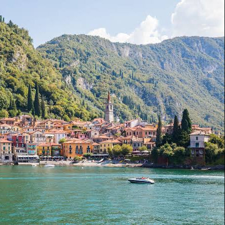A scenic view of a coastal town with colorful buildings, a church tower, and a small boat on turquoise water, backed by steep green mountains under a blue sky.