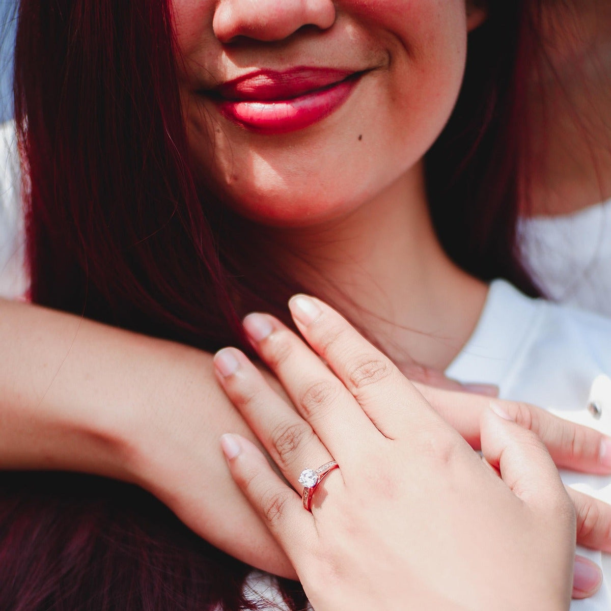 A close-up lifestyle shot of a woman wearing a delicate silver band ring with a small clear gemstone on her finger, with her hands crossed over her chest. She has dark auburn hair and is wearing a white top.
