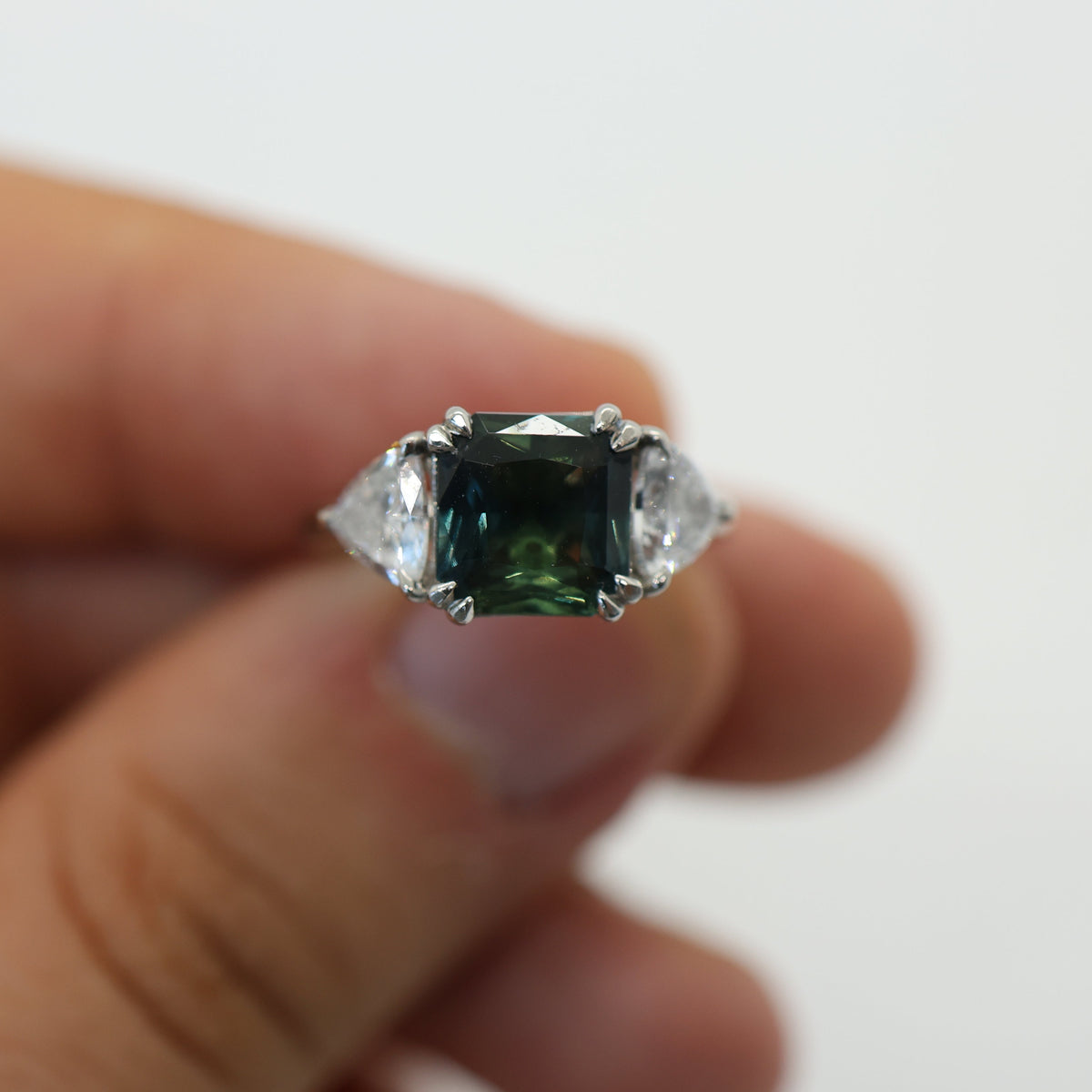 A hand holding a silver-toned ring featuring a square-cut dark green gemstone with two clear accent stones on either side, set against a plain white background.