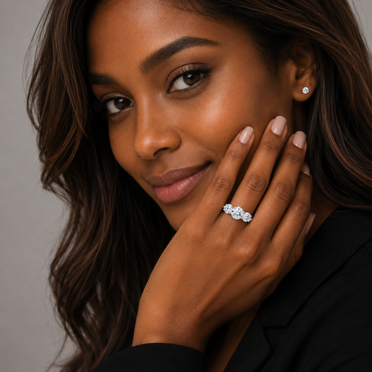 A close-up portrait of a woman wearing a sparkling diamond engagement ring on her finger, with a small diamond stud earring on her other ear. She is wearing a black blazer and posing with her hand near her face against a neutral gray background.