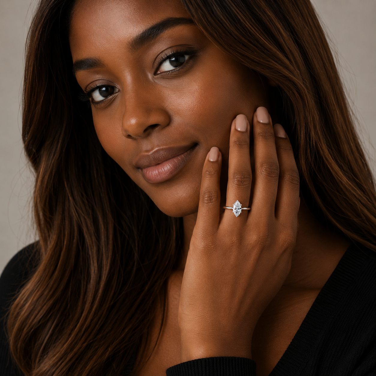 A close-up portrait of a woman with long brown hair wearing a diamond marquise-cut ring on her finger, with her hand resting against her cheek.