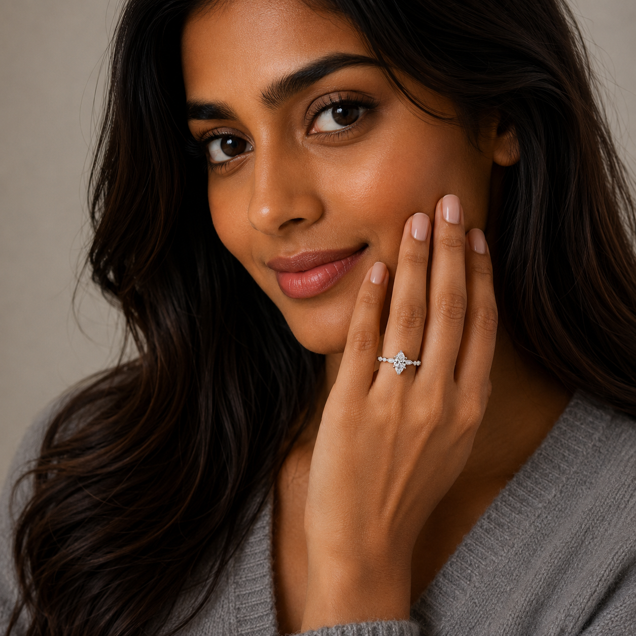 A close-up lifestyle shot of a woman with long dark hair wearing a silver ring with a clear marquise-cut gemstone on her ring finger, posing with her hand near her face against a soft beige background.
