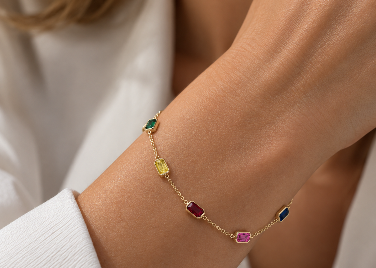 A delicate gold chain bracelet with five small, colorful rectangular gemstones (green, yellow, red, pink, blue) worn on a woman's wrist, styled with a white textured top.