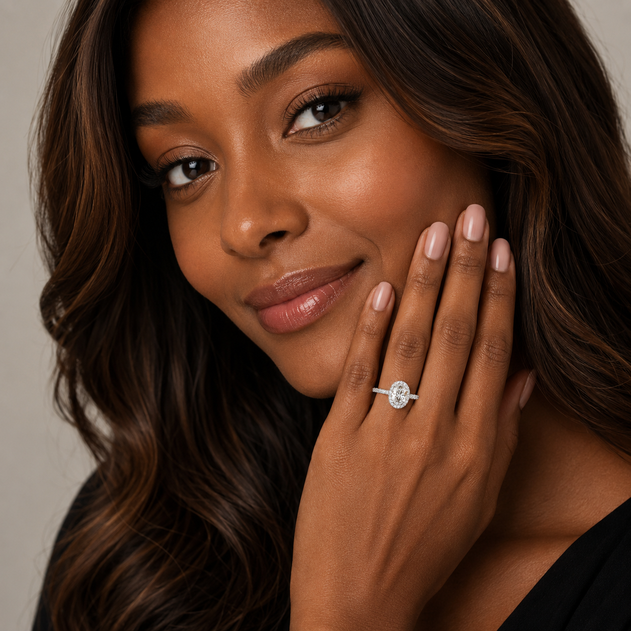 A close-up lifestyle shot of a woman with wavy brown hair wearing a silver engagement ring with a round-cut clear gemstone on her finger, with her hand resting against her cheek.
