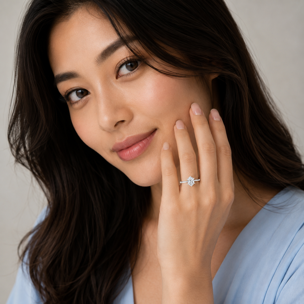 A close-up lifestyle shot of a woman with long dark hair wearing a delicate silver ring with a clear round-cut gemstone on her finger, set against a soft beige background.