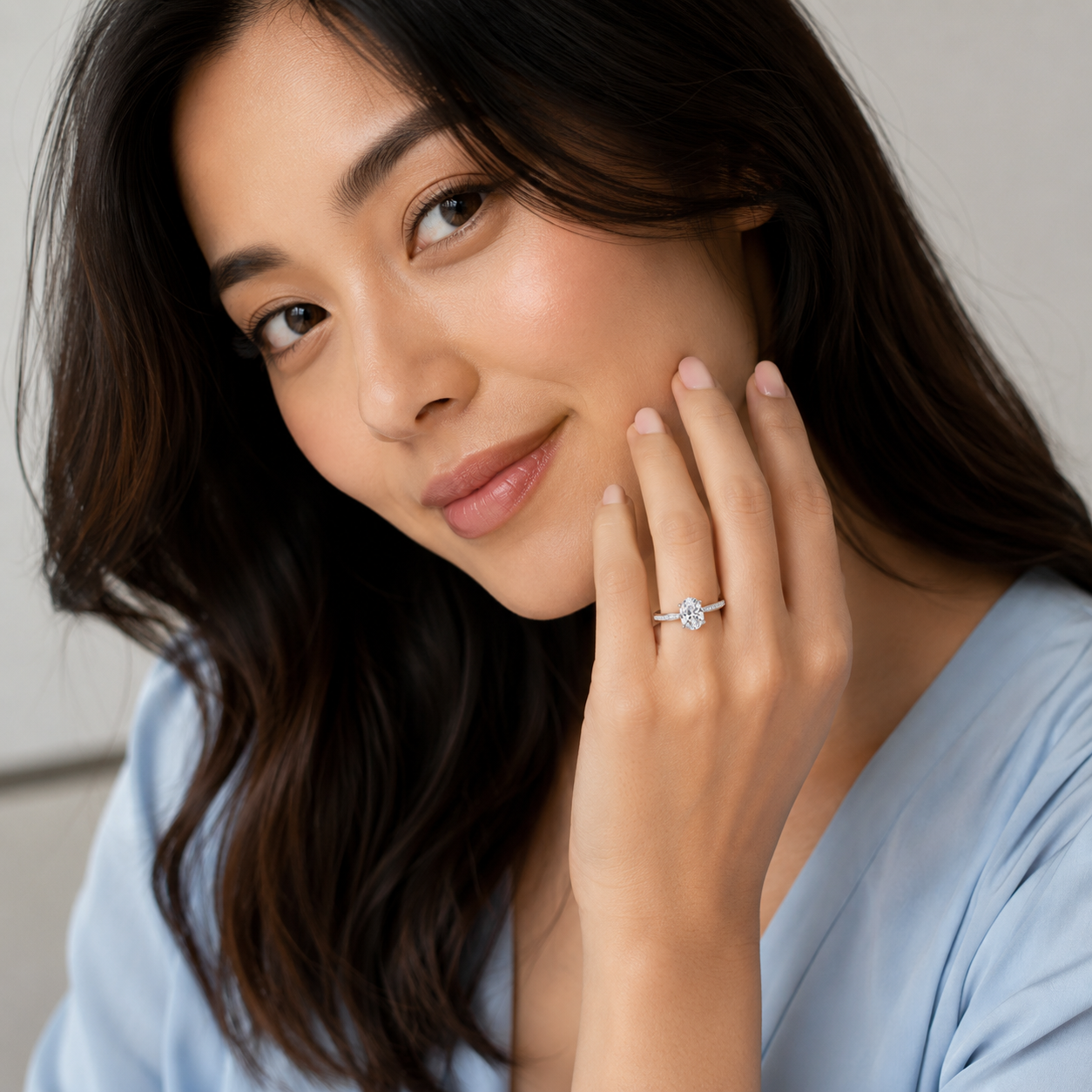 A close-up lifestyle shot of a woman with long dark brown hair wearing a delicate silver ring with a clear round-cut gemstone on her finger, set against a soft, light gray background.