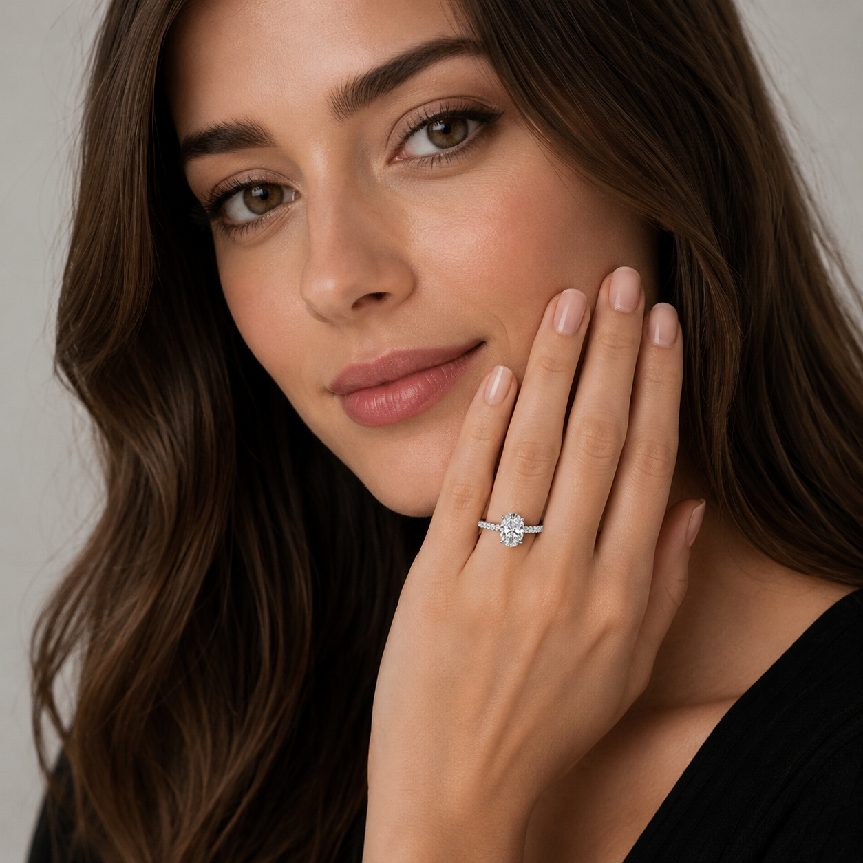 A close-up lifestyle shot of a woman with long brown hair wearing a delicate silver ring with a round-cut clear gemstone on her finger, posing with her hand near her face against a soft gray background.