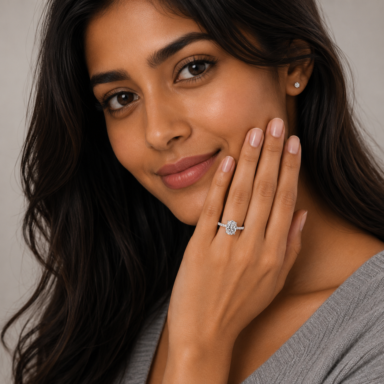 A close-up lifestyle shot of a woman with long dark hair wearing a silver-toned diamond engagement ring on her ring finger, with a small matching stud earring on her ear. She is smiling gently and touching her face with her hand, wearing a gray top against a soft neutral background.