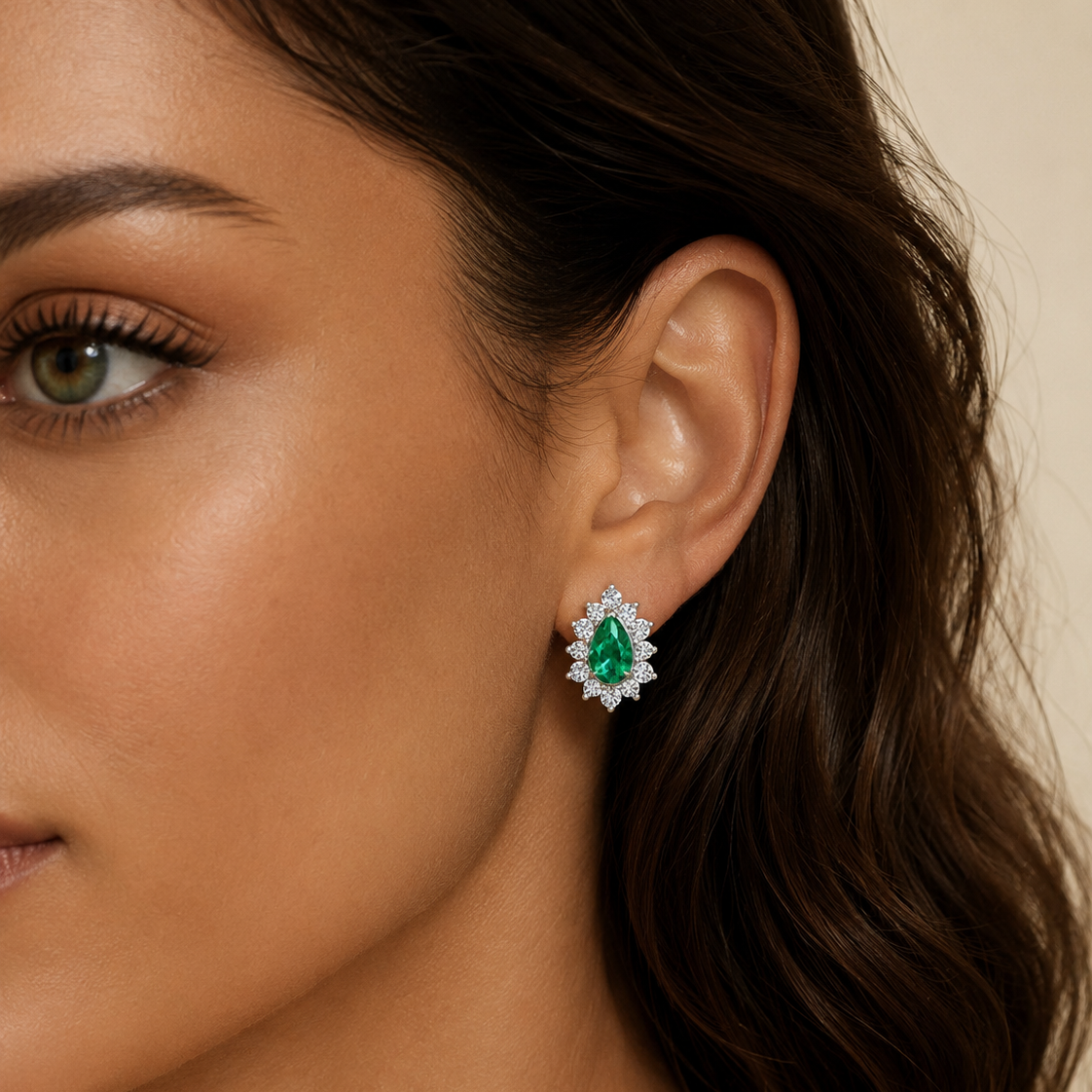 A close-up of a woman's ear wearing a teardrop-shaped stud earring with a green center stone surrounded by small clear stones, set against a soft beige background.