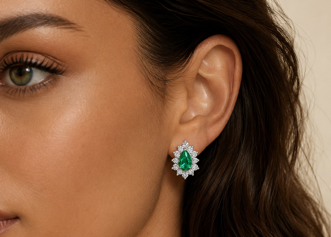 A close-up of a woman's ear wearing a teardrop-shaped stud earring with a green center stone surrounded by small clear stones, set against a soft beige background.