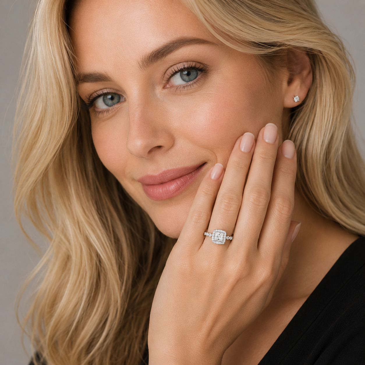 A close-up lifestyle shot of a blonde woman wearing a square-cut diamond engagement ring on her ring finger, with a small matching diamond stud earring on her ear. She is touching her cheek with her hand, which has manicured nails.