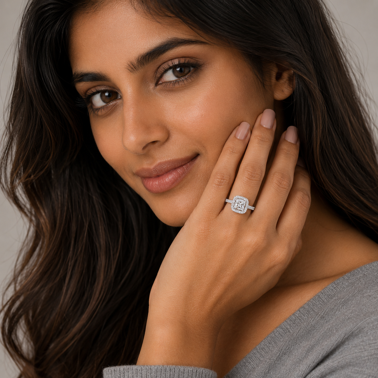 A close-up lifestyle shot of a woman with long dark brown hair wearing a square-cut diamond engagement ring on her finger, posing with her hand near her face against a soft neutral background.