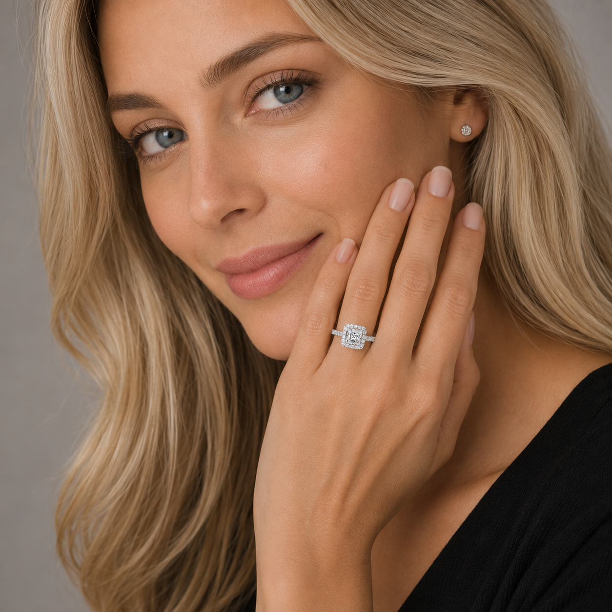 A close-up lifestyle shot of a woman with blonde hair wearing a square-cut diamond engagement ring on her finger and a matching stud earring, set against a neutral gray background.