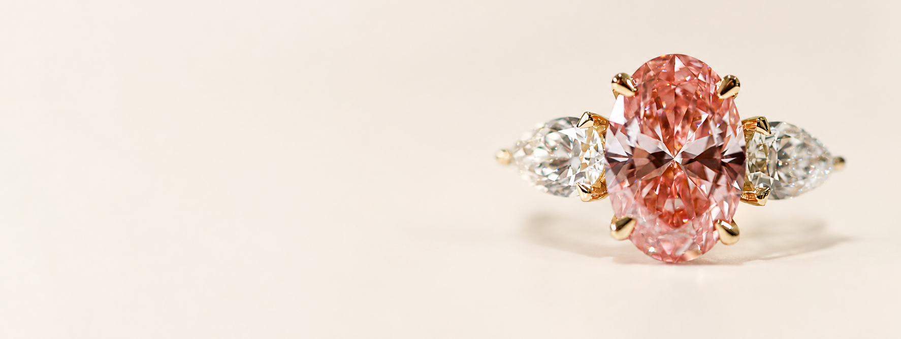 An oval pink diamond ring with yellow gold prongs, flanked by two marquise-cut clear diamonds on a warm cream background.