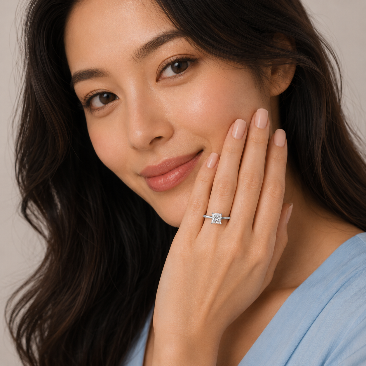 A close-up lifestyle shot of a woman with dark hair wearing a silver band ring with a square-cut clear gemstone, touching her cheek with her hand.