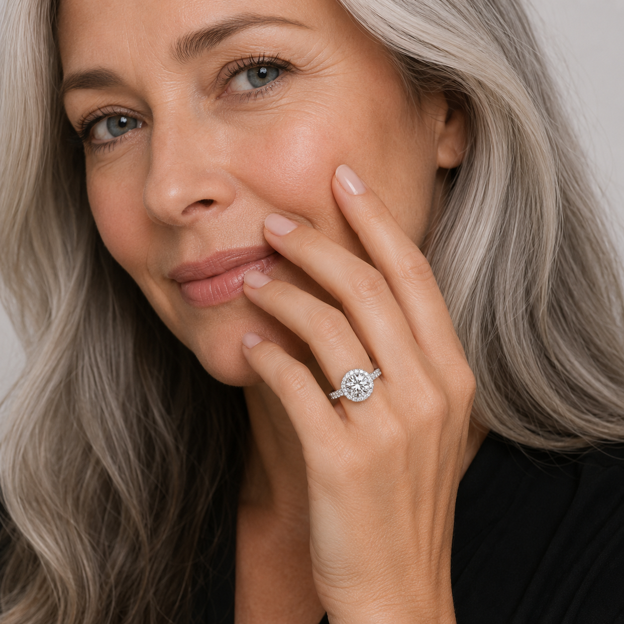 A close-up lifestyle shot of a woman with gray hair wearing a sparkling diamond engagement ring on her finger, with her hand resting near her face.