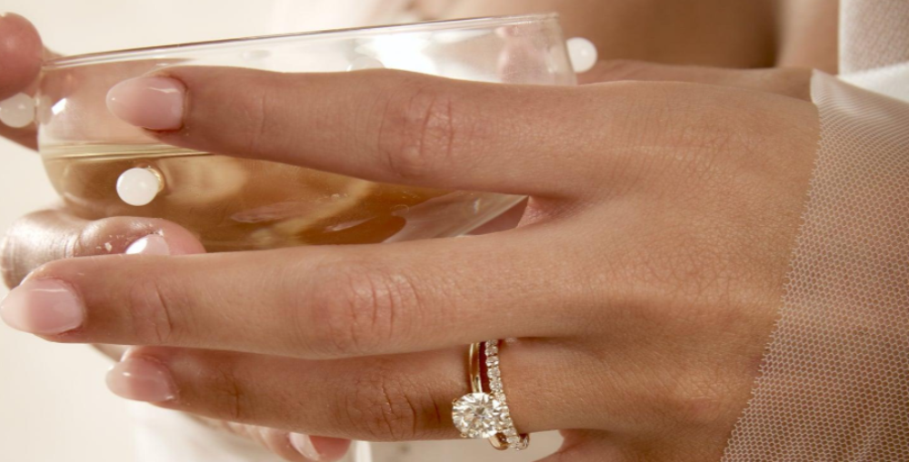 A close-up of a hand wearing a gold ring with a round-cut clear gemstone and small side stones, while holding a glass of champagne.