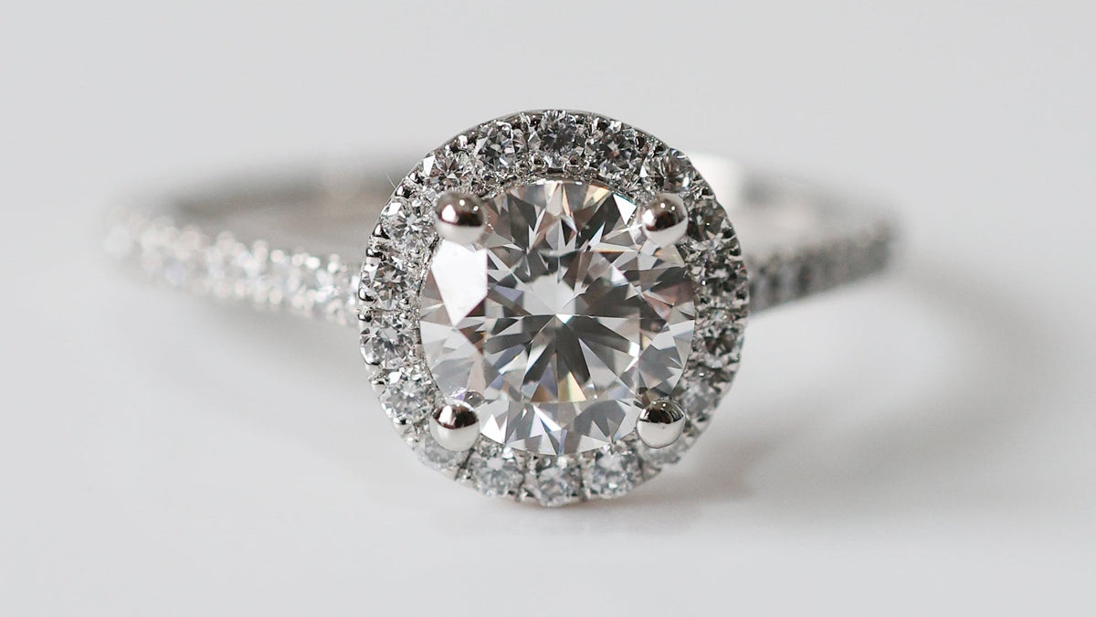 A round halo engagement ring featuring a large round-cut center stone surrounded by a double halo of smaller stones, set on a silver-toned band with pavé accents, photographed on a clean light gray background.