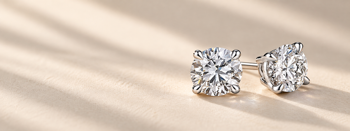 A pair of round brilliant-cut diamond stud earrings in a silver four-prong setting, resting on a warm beige textured surface with soft diagonal shadows.