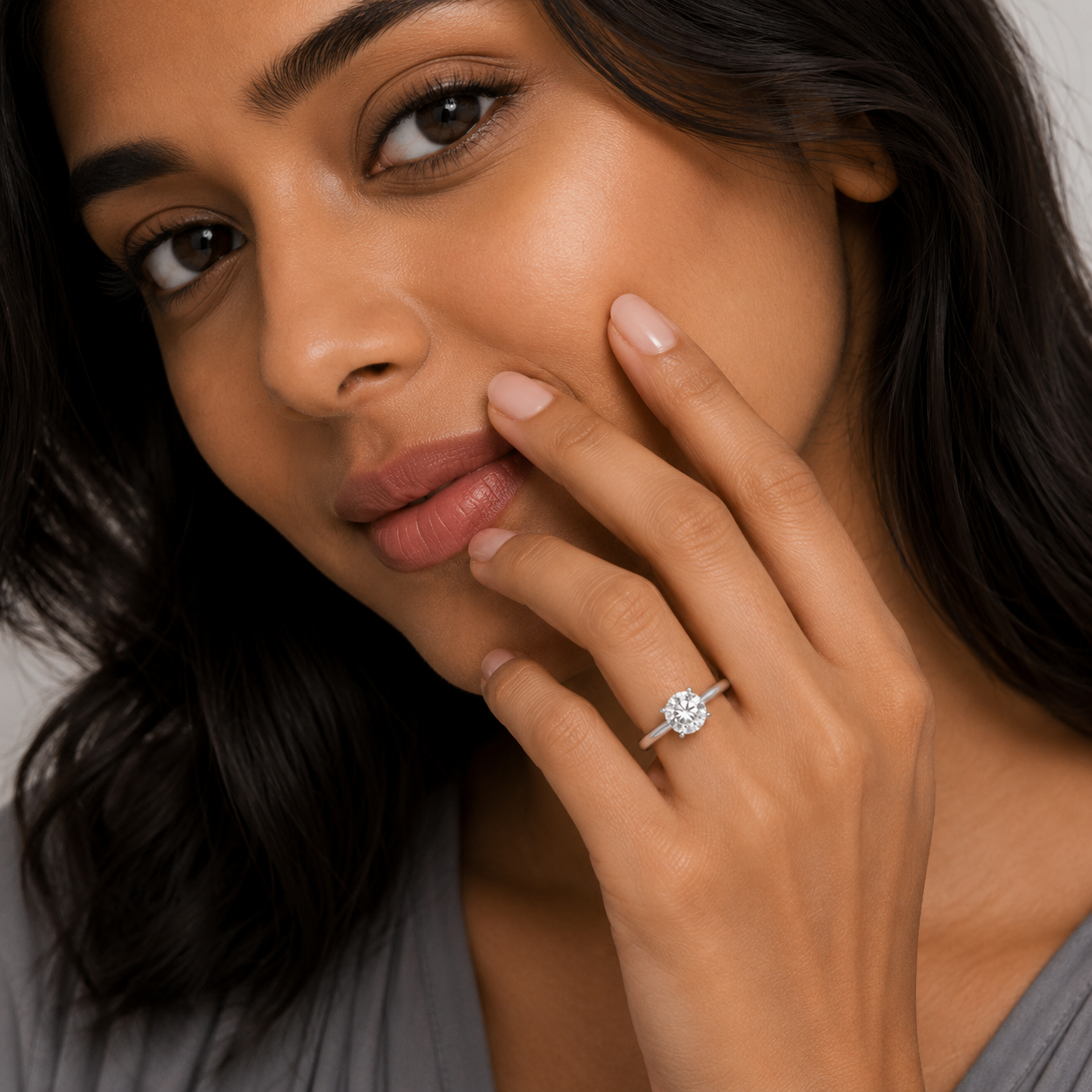 A close-up lifestyle shot of a woman wearing a silver-toned diamond engagement ring on her finger, with her hand resting against her cheek.