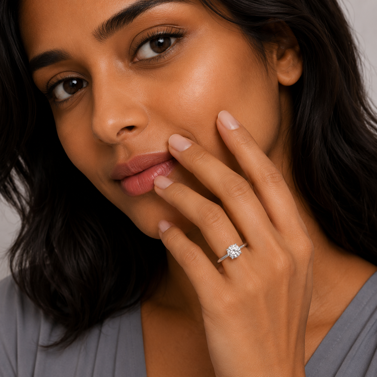 A close-up lifestyle shot of a woman wearing a single diamond ring on her ring finger. The ring features a round brilliant-cut center stone set in a silver-toned halo, with small accent stones around it.