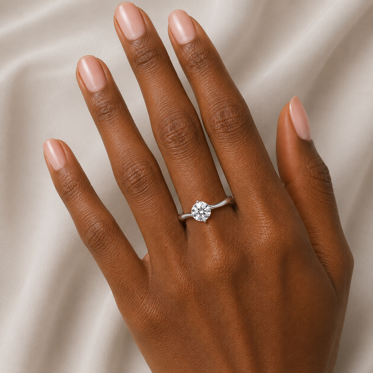 A close-up of a hand with manicured nails wearing a silver ring with a round clear gemstone, set against a soft beige satin fabric background.