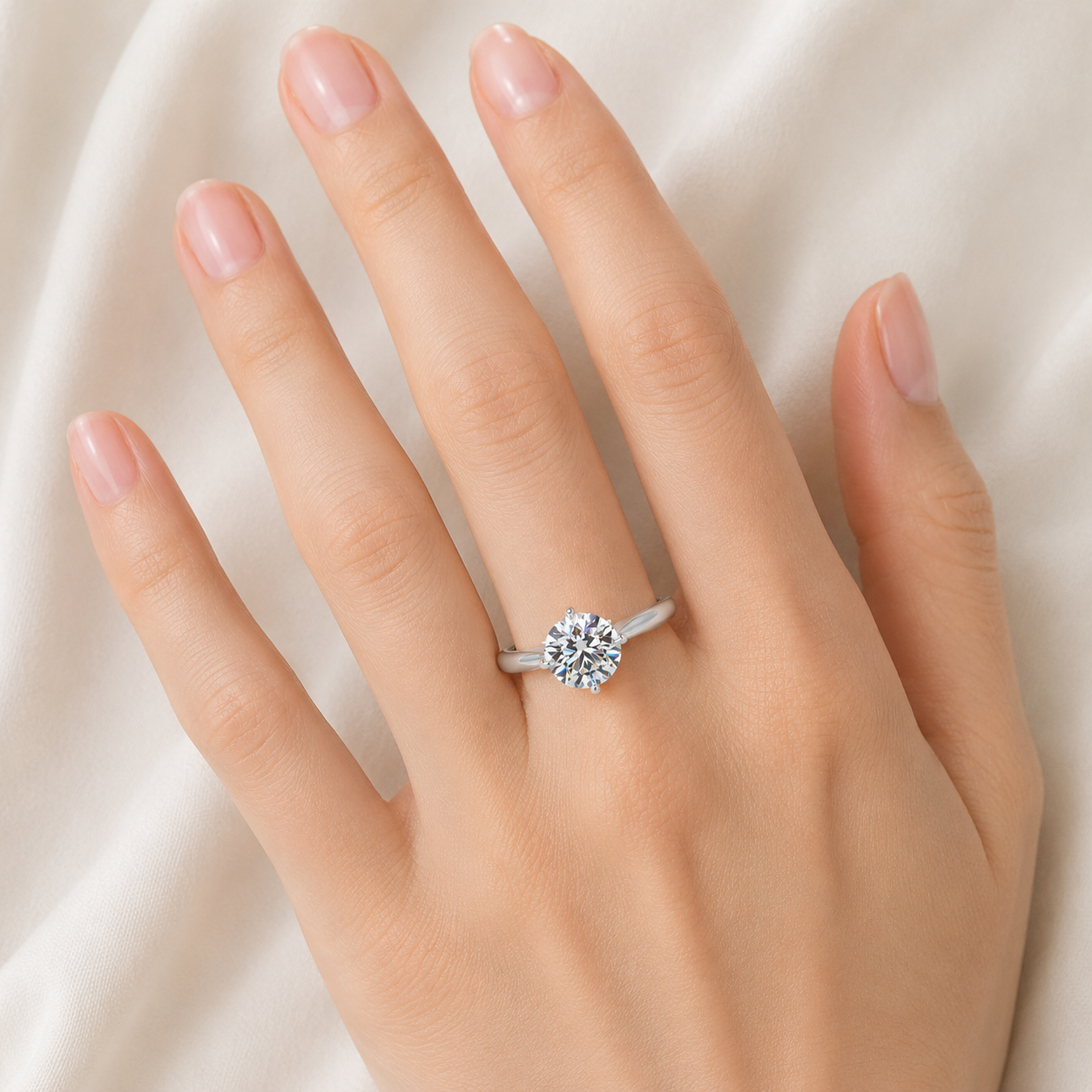 A silver-toned solitaire ring with a round clear gemstone, shown worn on a hand against a soft white fabric background.