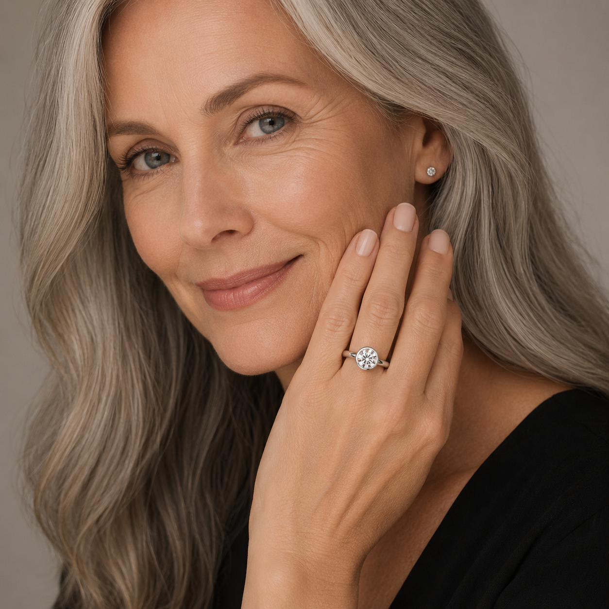 A close-up portrait of a woman with gray hair wearing a silver-toned ring with a round clear gemstone on her finger, against a soft neutral background.