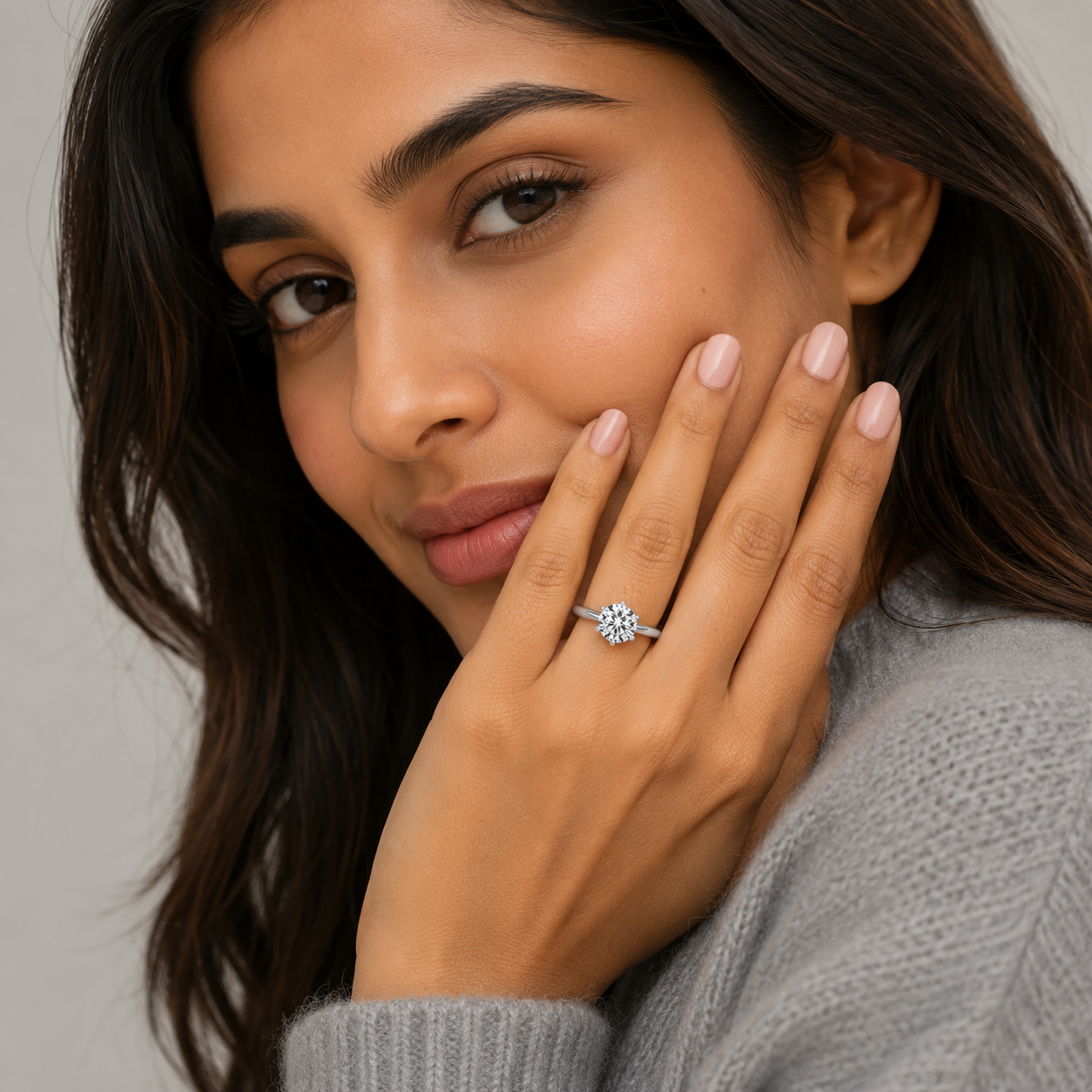 A close-up lifestyle shot of a woman wearing a silver ring with a clear round-cut gemstone on her finger, posing with her hand near her face against a soft gray background.