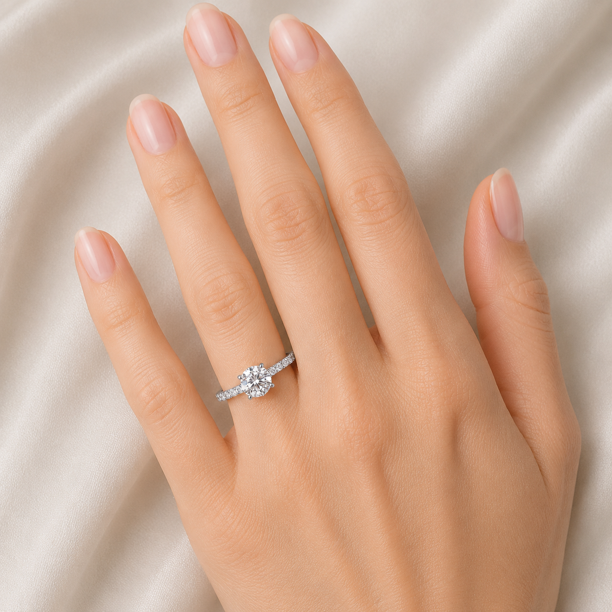 A close-up of a hand wearing a silver-toned ring with a round-cut center stone and small side stones, set against a soft beige satin fabric background.