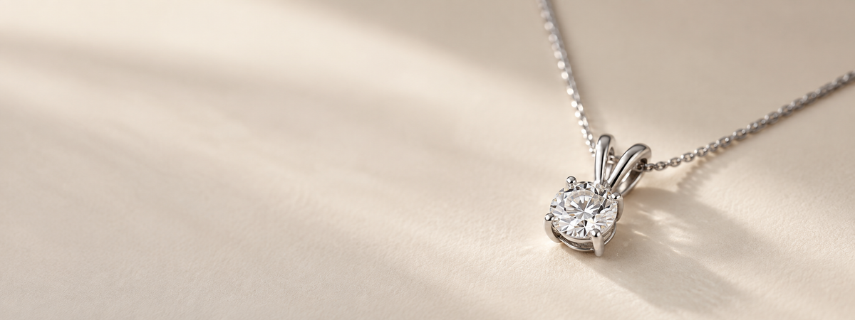 A silver-toned diamond pendant necklace on a delicate chain, resting on a warm beige textured surface with soft diagonal shadows.